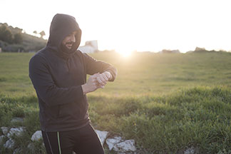 Pánské mikiny na sport: Muž v černé mikině s kapucí a v joggingových kalhotách je uprostřed krajiny. Má kapuci přes hlavu a dívá se na své hodinky na zápěstí.