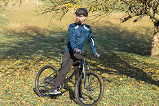 Vousatý cyklista s funkčními joggingovými kalhotami, mikinou, helmou a slunečními brýlemi stojí za slunečného dne na stráni a vychutnává si hezký okamžik.