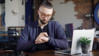 Pánské pletené svetry do kanceláře: Mladý muž s brýlemi a tmavě modrým kardiganem sedí v kavárně, laptop má na stole a prohlíží si své chytré hodinky.