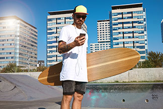 Skateboardista se slunečními brýlemi a kšiltovkou v triku oversized stojí ve skateparku. V jedné ruce má prkno, ve druhé mobilní telefon, na který se právě dívá.