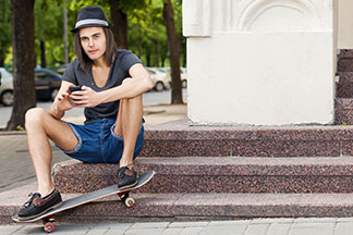 Stylově oblečený skateboardista s tmavým kloboučkem a džínovými šortkami sedí uvolněně na mramorových schodech a prohlíží si svůj mobil.