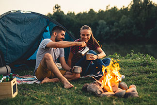Správné vybavení: Mladý pár v outdoorovém oblečení sedí před stanem u ohně, pije kávu a má dobrou náladu.