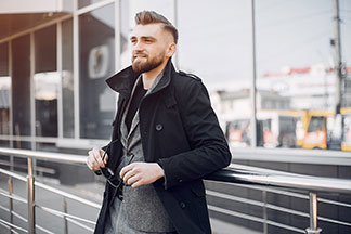 Pánská bunda do kanceláře: Mladý muž v krátkém černém kabátě a šedém saku se opírá o zábradlí a drží v ruce sluneční brýle.