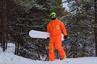 Snowboardista v oranžové lyžařské kombinéze běží mimo sjezdovku zasněženým zimním lesem.